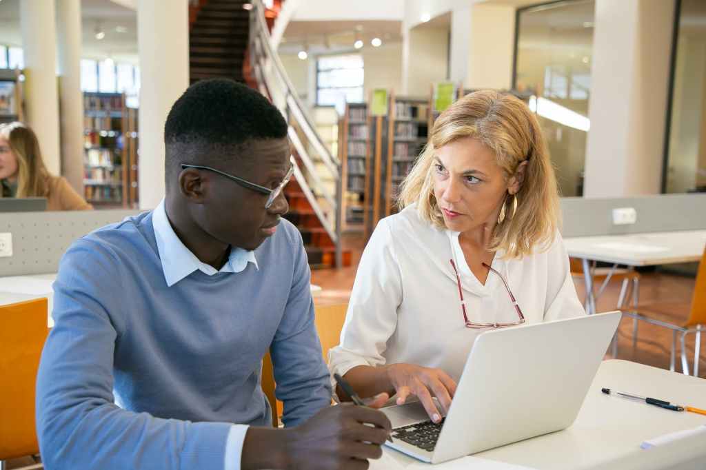 young black guy using laptop during exam preparation with female tutor in library